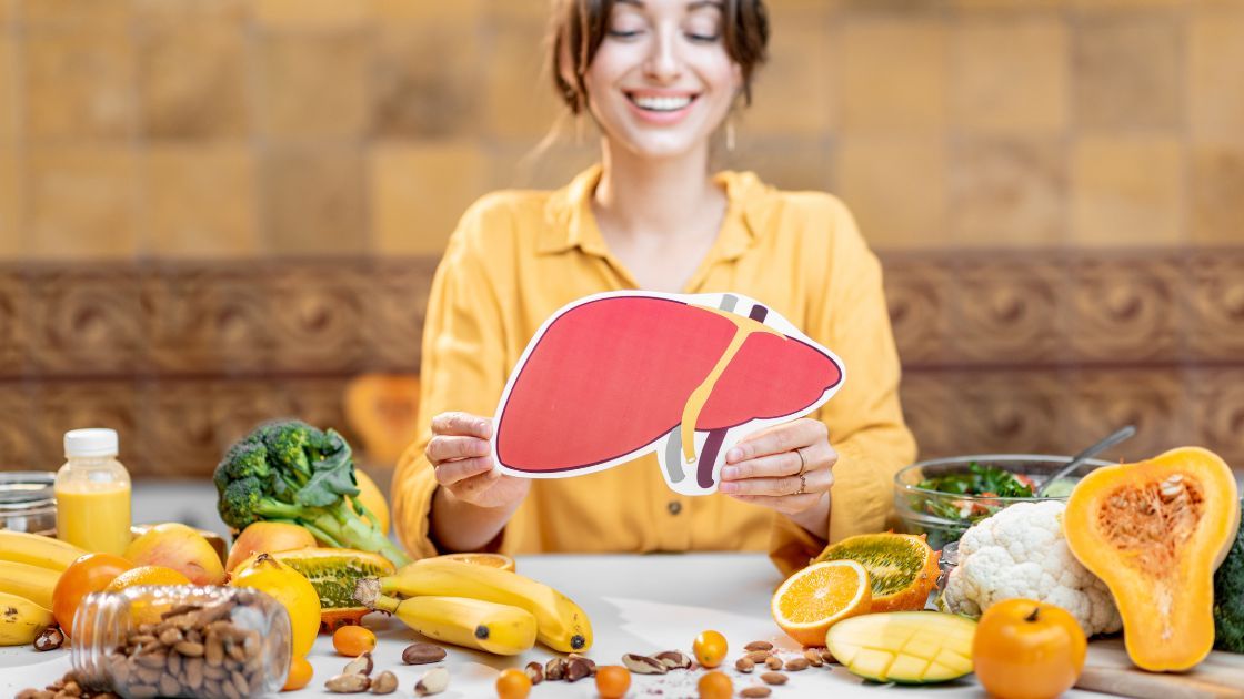 A smiling lady in a yellow shirt holding a liver-shaped paper with nutritious foods on the table.