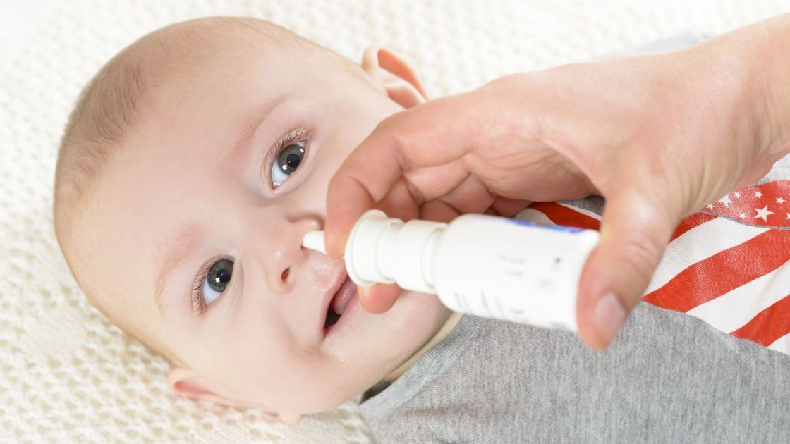 Cute baby, a hand seen, holding a nasal spray bottle, gently spraying on baby's left nostril.